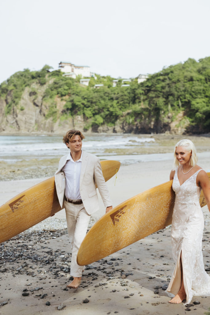 bride and groom walking with surf boards on beach
