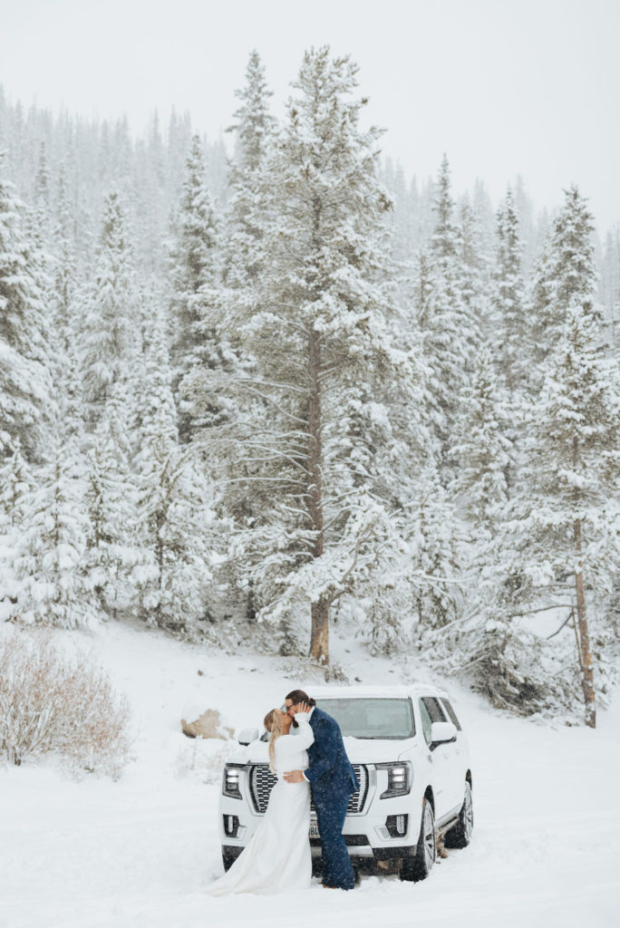 bride and groom kissing in front of car and snowy trees