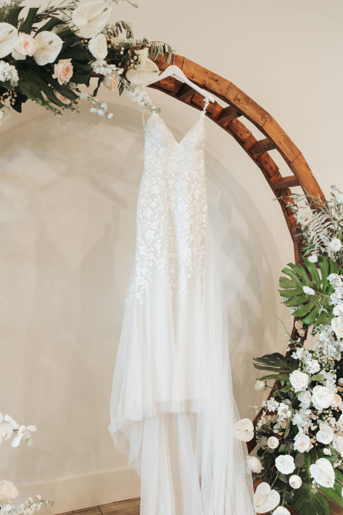 wedding dress hanging from wooden arch with white flowers and greenery