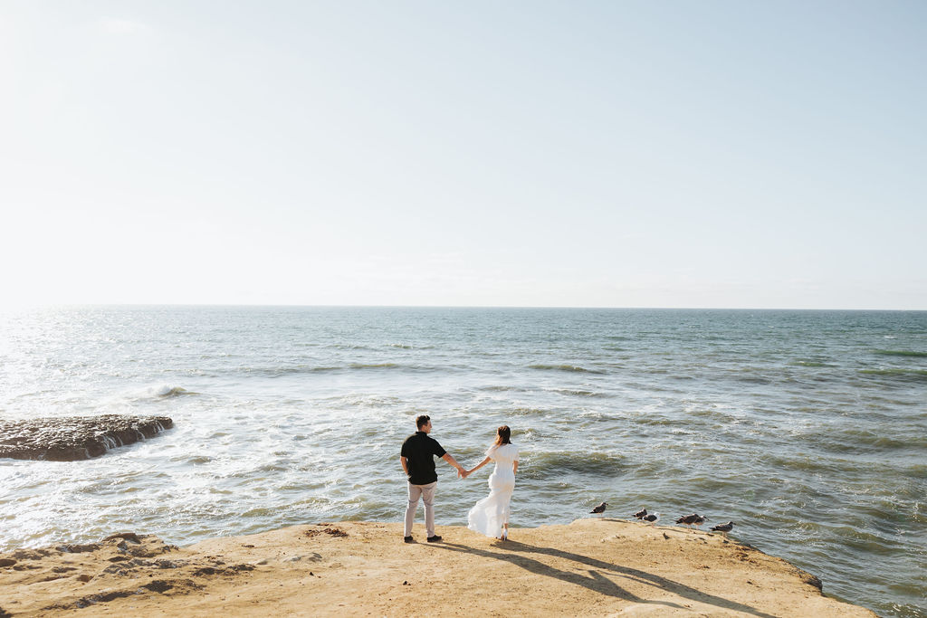engaged couple walking towards water on san diego cliff at sunset