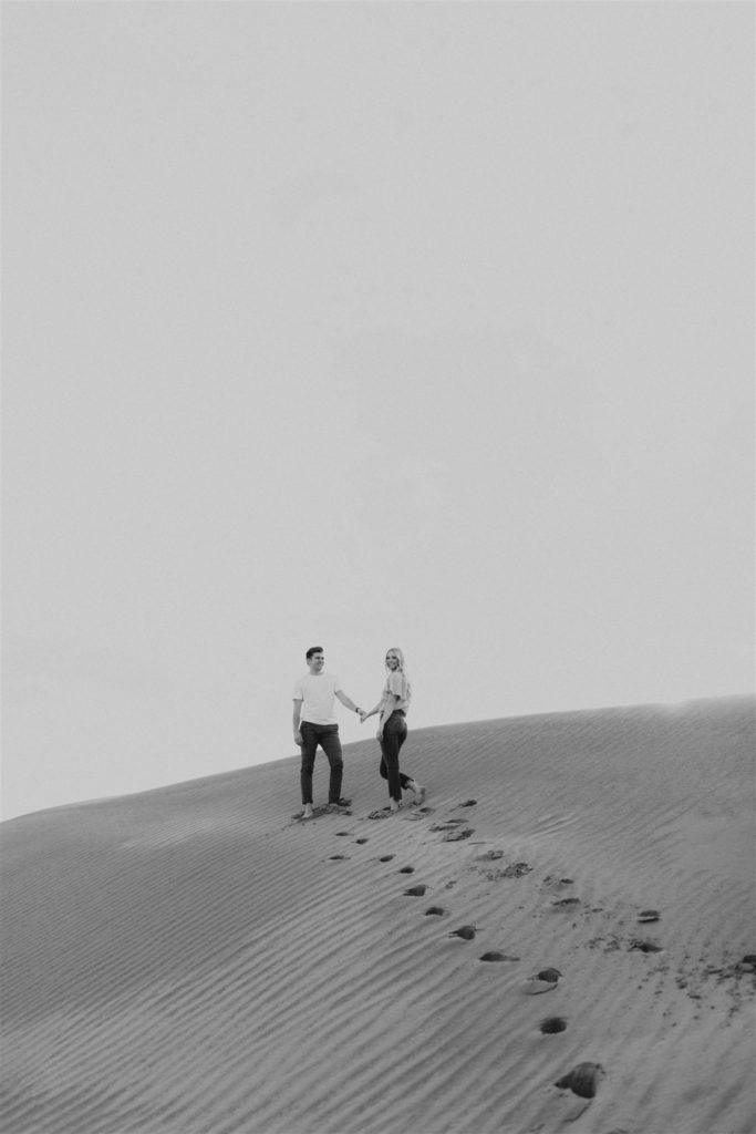 Black and white image of a Couple walking on a sand dune for their engagement session