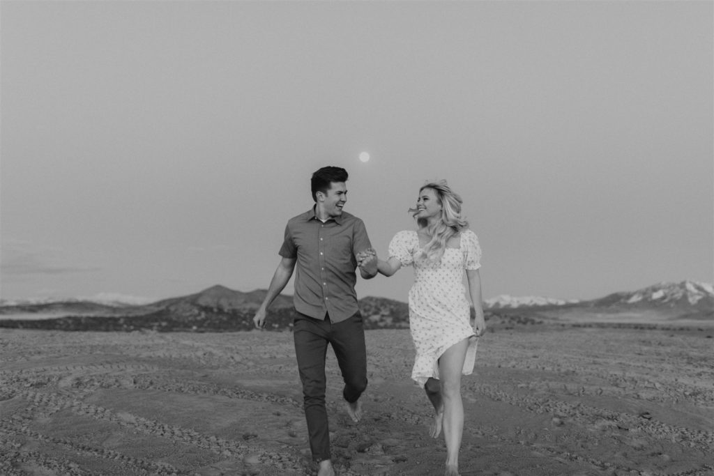 Couple running on sand dune in front of mountains for their engagement session