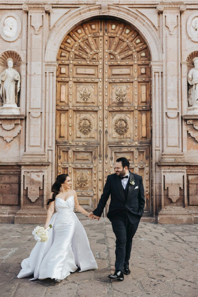 bride and groom holding hands and walking in front of large doors of cathedral