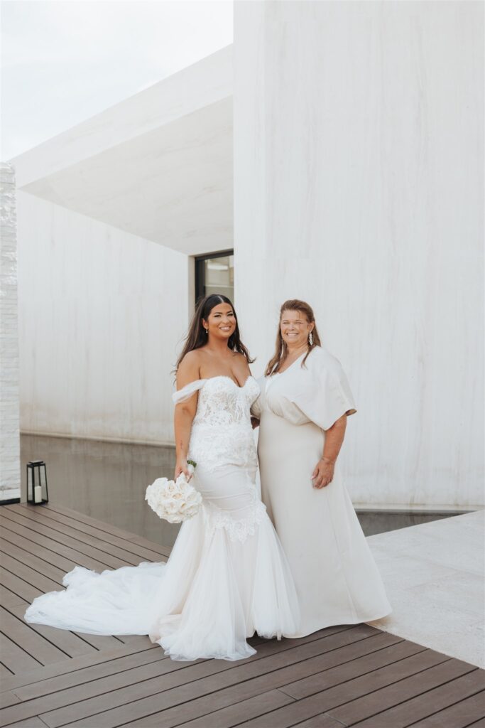 bride and mother posing in front of white wall with white flowers