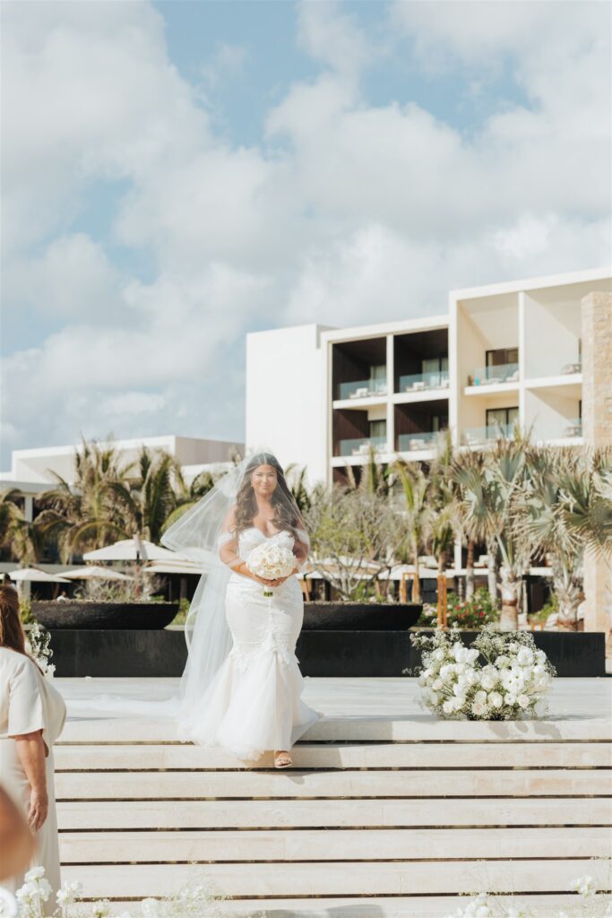 bride walking down the aisle for wedding ceremony with floral arch at the beach in los cabos